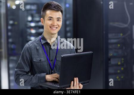 Smiling Chinese male technician using laptop standing in computer room Stock Photo