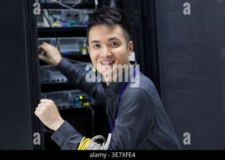 Smiling Chinese male technician with roll of cables on arm doing maintenance in computer room Stock Photo