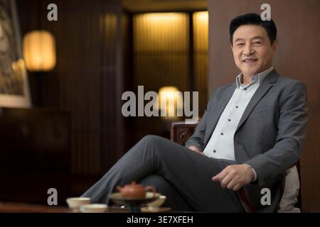 Cheerful Chinese businessman wearing gray suit sitting with legs crossed on chair in office Stock Photo