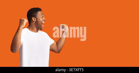 Excited Black man wearing white shirt with clenched fists standing against orange background Stock Photo