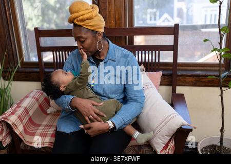 Black mother wearing yellow head wrap and long sleeve denim shirt carrying and playing with her baby boy with pacifier while sitting on wooden sofa... Stock Photo