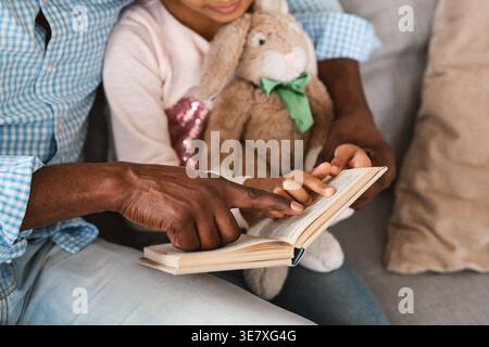 Cropped image of Black grandfather and granddaughter pointing at book while reading together on sofa at home Stock Photo