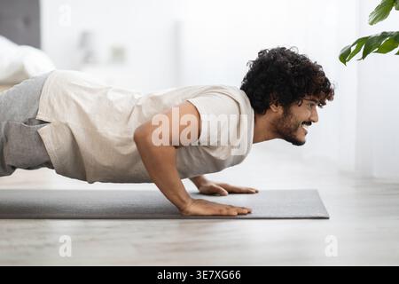 Asian man with curly hair looking away while planking on yoga mat in bedroom at home Stock Photo