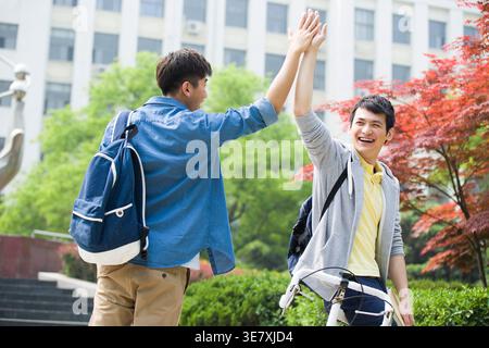 Smiling Chinese male college students giving high five to each other while standing on campus during daytime Stock Photo