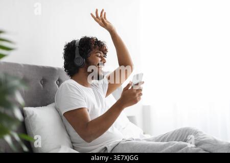 Bearded Indian man wearing headphones waving hand having video call on smartphone sitting on bed in bedroom at home Stock Photo