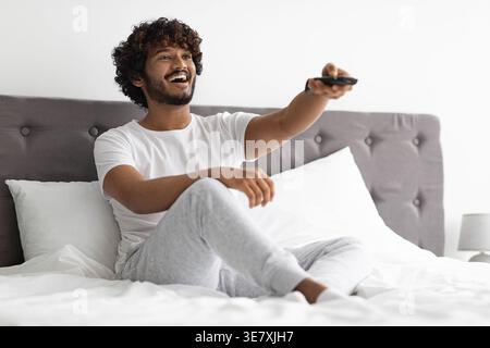 Bearded Indian man with curly hair using remote control while watching TV sitting on bed in bedroom at home Stock Photo