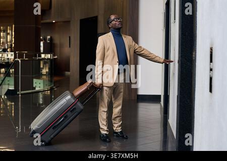 Black businessman with luggage wearing eyeglasses looking up pressing button for elevator while standing on hallway in hotel Stock Photo
