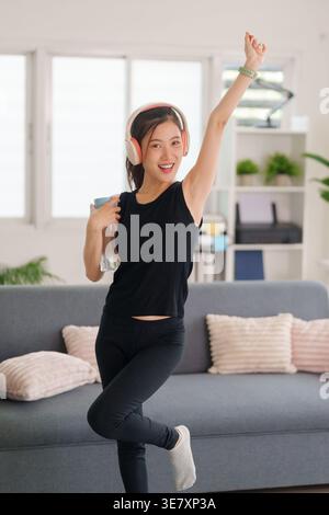 Cheerful Asian woman enjoys active home workout listening to music. She holds a water bottle expressing pure joy. Stock Photo