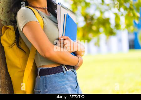Asian female student with backpack holding books leaning against tree trunk in university during daytime Stock Photo