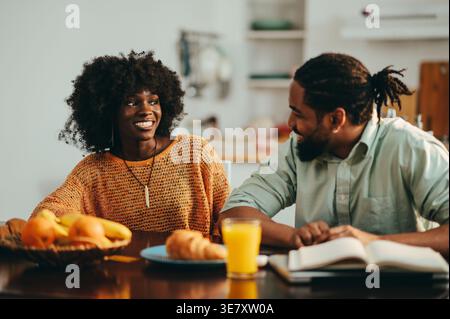 Smiling Black couple talking while sitting at table with foods, book, and laptop during breakfast at home Stock Photo