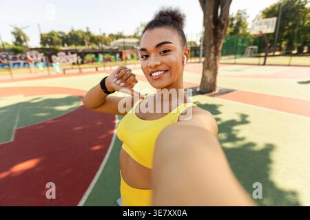 Black female in fitness wear pointing thumb at herself standing on basketball court on sunny day Stock Photo