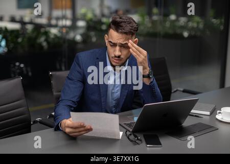 Stressed Arab businessman reading document and touching his temple while sitting at desk with laptop and smartphone in office Stock Photo