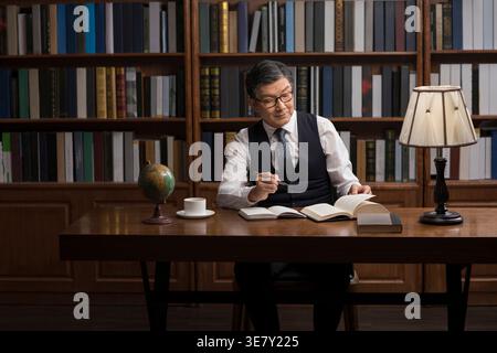 Focused Chinese businessman in eyeglasses reading book studying and drinking coffee while sitting at table in his home library Stock Photo