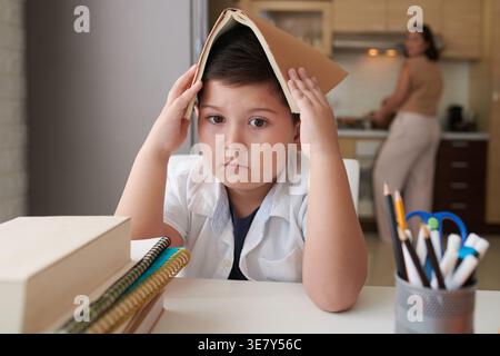 Sad Mixed race son covering head with books sitting against her Asian mother in kitchen at home during daytime Stock Photo