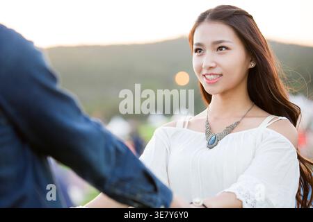 Smiling Chinese woman holding hands with boyfriend having fun while standing on concert ground with tents at music festival Stock Photo