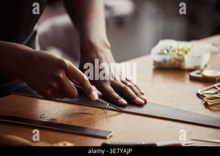 Hands of Black woman artisan cutting leather creating handmade leather belt with tool while standing in workshop Stock Photo