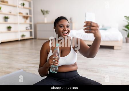 Curvy Black woman in wireless earphones holding bottle of water taking selfie using smartphone while sitting on yoga mat in living room at home Stock Photo