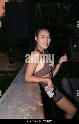 A young Thai woman in brown athletic gear smiles while leaning on a wooden railing during evening, holding a smartphone after a training session to re Stock Photo