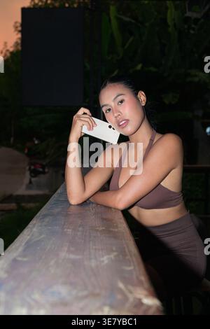 A young Thai woman in brown activewear leans on a wooden railing at an outdoor terrace during twilight, holding her mobile phone while looking thought Stock Photo
