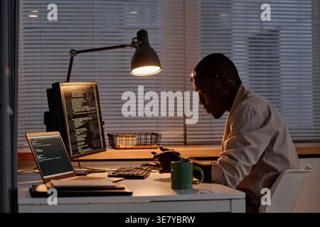 Focused Black male programmer in eyeglasses examining computer codes working overtime in office Stock Photo