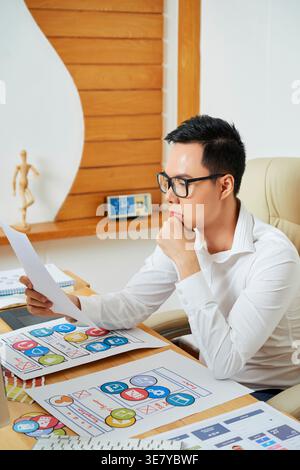 Asian male graphic designer holding paper while looking it sitting on chair in office Stock Photo