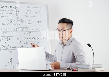 Serious Asian male math teacher pointing at whiteboard with formulas while explaining topic to students sitting at desk during online class in his ... Stock Photo