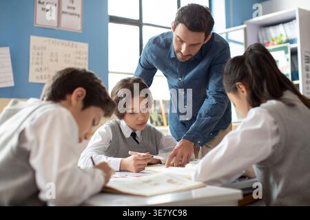 Diverse group of students doing school work while caucasian male teacher assisting them in classroom Stock Photo
