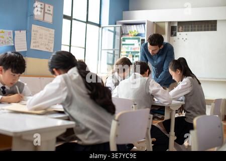 Diverse group of students doing school work while caucasian male teacher assisting them in classroom Stock Photo