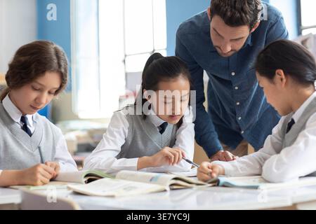 Diverse group of students doing school work with caucasian male teacher assisting them in classroom Stock Photo