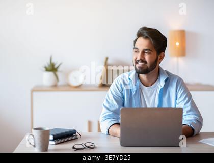 Smiling Middle Eastern man looking away working on laptop sitting at table at home office Stock Photo