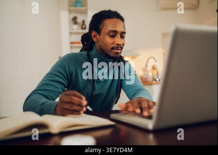 Serious Mixed race man with locks holding pen taking notes on notebook sitting at table with laptop at home Stock Photo
