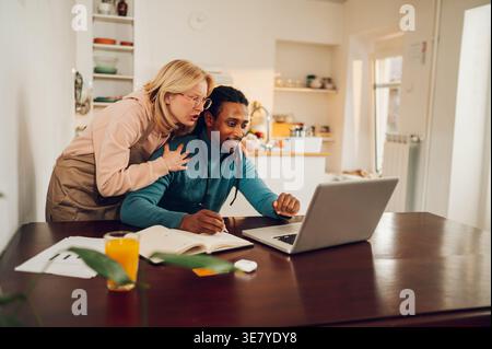 Smiling Mixed race man with locks using laptop sitting at table near caucasian girlfriend hugging him from behind at home Stock Photo