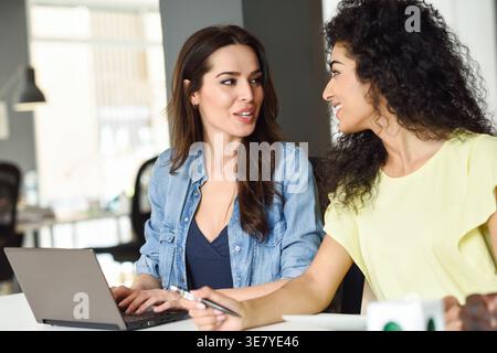 Smiling Middle Eastern woman with curly hair talking to caucasian female colleague typing on laptop while sitting at table in the office Stock Photo