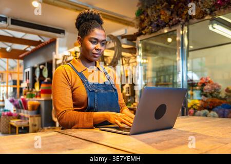 Hispanic female employee managing orders using laptop while standing at wooden table in flower shop Stock Photo