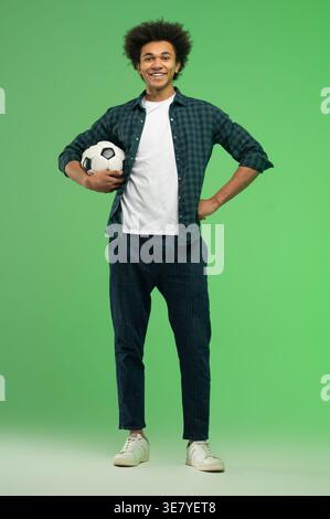 Smiling Black man with afro hair holding soccer ball standing against green background in studio Stock Photo