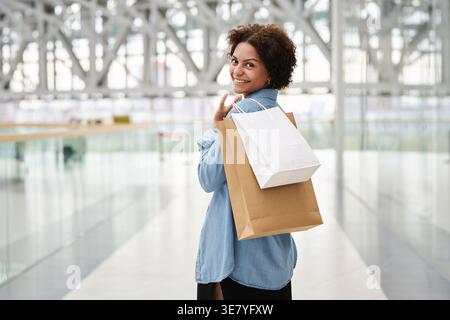 Black female teenager walking around looking back while holding paper bags in mall hallway Stock Photo