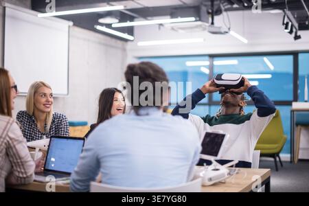 Smiling Black male developer with braids wearing VR headset and experiencing virtual reality while sitting at table with diverse colleagues laughin... Stock Photo