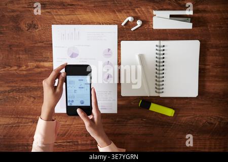 Female designer using smartphone while sitting at table with notebook and documents in office Stock Photo