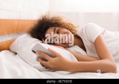 Smiling Black woman with curly having video call using smartphone lying on bed in bedroom at home Stock Photo