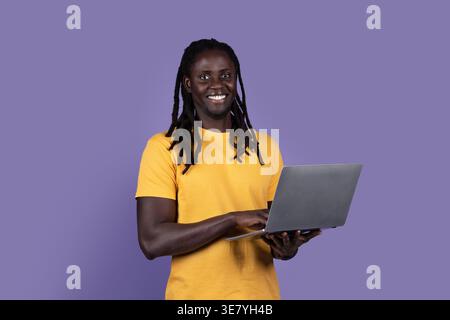 Smiling Black man with dreads wearing yellow shirt using laptop while standing against purple background in studio Stock Photo