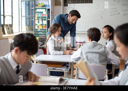 Diverse group of students doing school work while caucasian male teacher assisting them in classroom Stock Photo