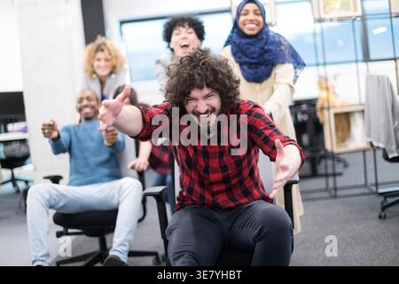 Cheerful Mixed race male employee raising arms having fun with coworkers pushing him sitting on chair in office Stock Photo
