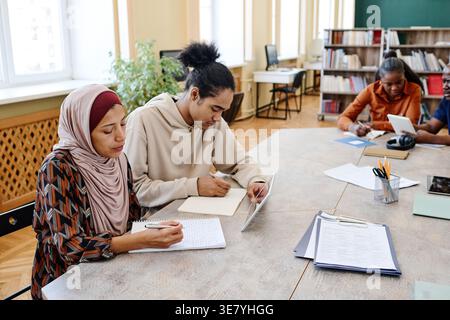 Hispanic Muslim female and Black male immigrants sitting at table using tablet together and taking notes attending class in library Stock Photo