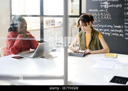 Mixed race female student using tablet sitting at table with Black female teacher leaning chin on hands and looking at laptop in classroom Stock Photo