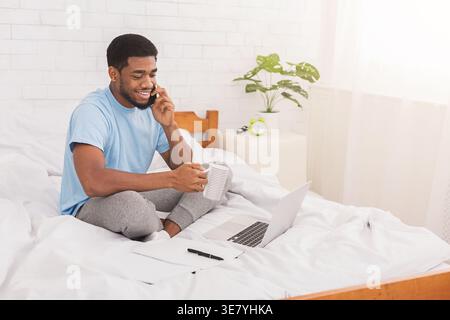 Smiling Black man having phone call and holding mug while sitting on bed with laptop at home Stock Photo
