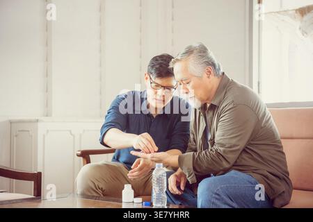 Asian middle-aged man giving medicine to his senior father sitting beside him on couch at home Stock Photo