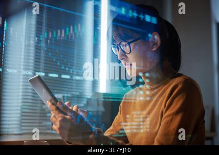 Focused Asian business woman wearing eyeglasses using tablet projecting the program sitting at table in office at night Stock Photo