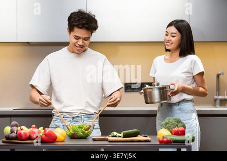 Asian man mixing vegetables in glass bowl preparing salad next to wife holding cooking pot standing near table in kitchen at home Stock Photo