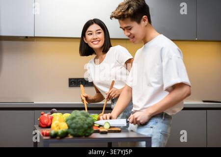 Asian woman mixing vegetables in bowl looking at husband slicing cucumber while standing at table in kitchen at home Stock Photo
