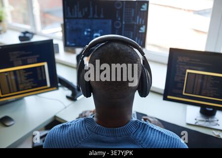 Back view of Black male programmer wearing headphones while typing codes on computer sitting at table in office Stock Photo